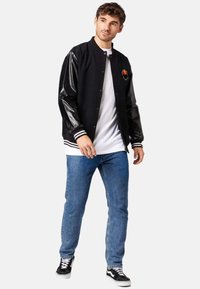 Black varsity jacket with leather sleeves, ribbed cuffs, and collar. Worn over a white t-shirt, paired with blue jeans and black sneakers.