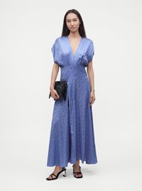 Femme portant une robe bleue à motifs avec un col en V profond, tenant une pochette noire et portant des sandales à talons noires sur fond blanc.