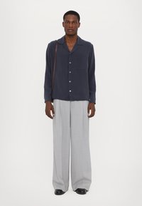 Marineblaues Hemd mit Knöpfen und einer Brusttasche, kombiniert mit weit geschnittenen, hellgrauen Hosen. Schwarze Schuhe vervollständigen das Outfit.