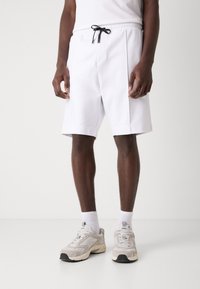 Shorts de sport blancs avec une taille à cordon de serrage, dotés de poches latérales et d'une coupe détendue, associés à des baskets claires et des chaussettes blanches.