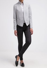 Blazer gris ajustado con un cuello alto y sin cierre, combinado con pantalones negros de corte slim y mocasines negros; tejido liso y diseño sencillo.