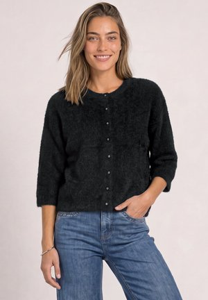 Femme souriante aux cheveux mi-longs portant un pull noir duveteux à boutons et un jean bleu, debout avec une main dans la poche.