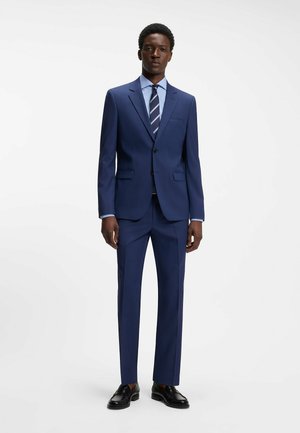 Homme portant un costume bleu marine sur mesure avec un pantalon assorti, une chemise bleu clair, une cravate rayée bleu marine et des chaussures habillées noires, debout devant un fond blanc.