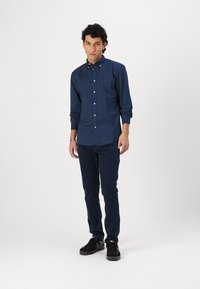 Navy button-up shirt with white polka dots and long sleeves, paired with dark blue jeans and black sneakers, standing against a white background.