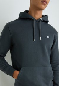 Katoenmix donkergrijze hoodie met een kangoeroezak, trekkoord en een klein wit logo op de borst. Soepele textuur en een getailleerd ontwerp.