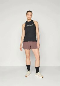 Črni brezrokavnik za šport z napisom "Normal" ter širokimi rjavimi kratkimi hlačami. Obleka je dopolnjena s svetlimi teniskami in črnimi nogavicami do gležnjev.