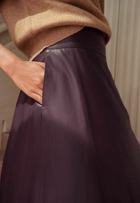 Falda de cuero púrpura con una textura suave y un diseño elegante. Presenta un bolsillo lateral y una cinturilla alta, combinada con un suéter marrón claro.