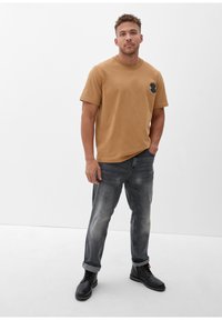 Lichtbruine katoenen t-shirt met een zwart berenlogo op de linkerkant van de borst, gecombineerd met vervaagde grijze denim jeans en zwarte veterschoenen.