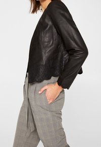 Chaqueta de cuero negra corta con dobladillo en forma de ola y detalles de corte intrincados, llevada sobre pantalones de cuadros grises con la cintura atada.