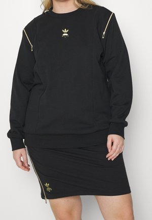 Persoon draagt een zwarte top met lange mouwen en rok met gouden ritsen en klein gouden Adidas-logo op de borst en zoom van de rok.