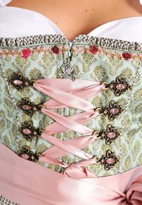 Gros plan sur un corset en brocart vert avec un laçage en ruban de satin rose et des fermoirs métalliques décoratifs ornés de pierres précieuses roses, porté par une personne vêtue d'un chemisier blanc.