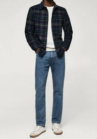 Zwart-blauwe geruite flanellen overhemd, witte ronde hals t-shirt, blauwe spijkerbroek en witte sneakers met tan accenten.