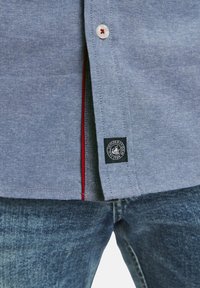 Helles blaues Button-Down-Hemd aus strukturiertem Stoff, mit einem roten Akzent entlang der Innenseite der Knopfleiste und einem Markenetikett am unteren Saum.