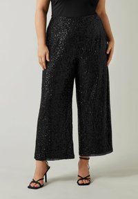 Pantalons larges noirs pailletés avec une surface texturée, présentant une taille haute et une coupe ample, accompagnés de talons noirs à brides.