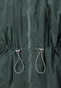 Tamnozelena jakna s teksturiranim materijalom, prednjim zipom i podesivom vezicom u struku s metalnim detaljima.