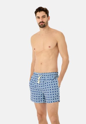 Homme torse nu aux cheveux foncés et à la barbe, portant un short de bain bleu et blanc à motifs avec des poches avant et un cordon blanc.
