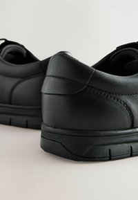 Chaussures en cuir noir avec une semelle en caoutchouc noir texturé, présentant une finition lisse et des coutures latérales, mettant en avant le talon et les détails arrière.
