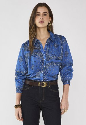 Vrouw met lang bruin haar die een blauw overhemd met patroon, een zwarte broek, een bruine riem, gouden oorringen en armbanden draagt.