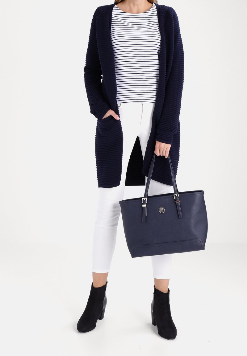 Un cardigan ouvert côtelé marine sur une chemise rayée, associé à un pantalon blanc et des bottines noires. Tenant un sac fourre-tout structuré marine.