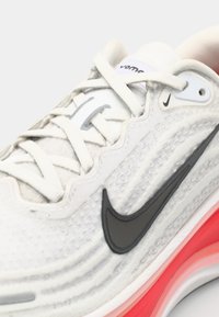 Baskets blanches avec une tige texturée, logo Swoosh noir de Nike, et une semelle dégradée rose. Présente des accents gris et des lacets plats.
