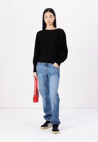 Zwarte gebreide trui met lange mouwen, gecombineerd met lichtblauwe baggy jeans en zwarte sneakers. Houdt een rode handtas vast.