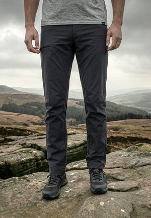 Bărbat stând pe teren stâncos, purtând pantofi negri de drumeție, pantaloni gri închis cu buzunar lateral cu fermoar și tricou gri deschis.