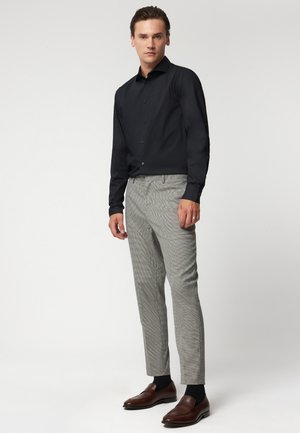 Czarna koszula z długim rękawem, szare spodnie w kratkę, brązowe mokasyny i czarne skarpetki. Strój jest dopasowany, o dobrze skrojonej sylwetce i subtelnych wzorach.