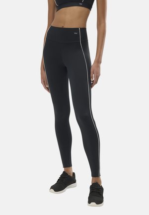 Mujer con leggings negros de cintura alta y franjas laterales blancas y zapatillas deportivas negras, de pie frente a un fondo liso.