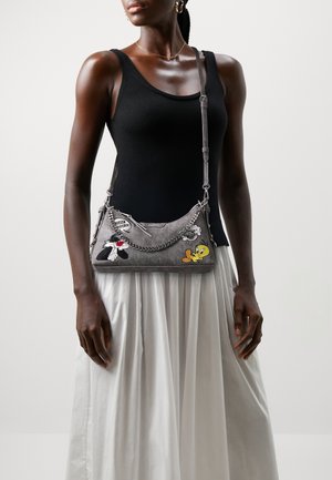 Kvinde iført sort ærmeløs top og hvid nederdel, bærer grå crossbody taske med Tweety og Sylvester tegneserie patches.