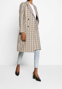 Abrigo cruzado en un patrón a cuadros amarillo, marino y blanco. Presenta solapas anchas y un corte largo y entallado. Combinado con jeans azul claro y tacones negros.