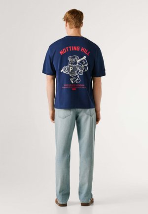 Majica od pamuka mornarsko plave boje s grafičkim dizajnom psa i natpisom "NOTTING HILL" u crvenoj boji na leđima. Uparena s trapericama svijetloplave boje.