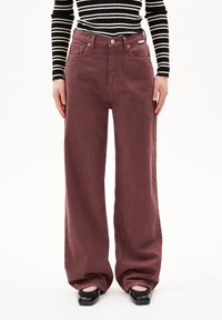 Weite Hose aus burgunderfarbenem Denim mit hoher Taille, fünf Taschen, Metallknopf und subtiler Textur. Kombiniert mit schwarzen Ballerinas.