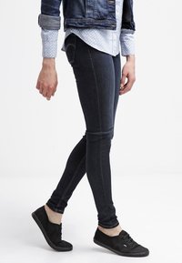 Persona vestida con jeans ajustados oscuros, zapatillas negras, una camisa clara con estampado y una chaqueta de mezclilla enrollada, de pie sobre un fondo blanco.