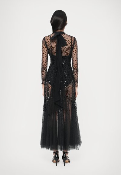 Black gown with long sheer sleeves, embellished with sequins. Features a flowing tulle skirt and a tied back detail. Glossy black heels visible.