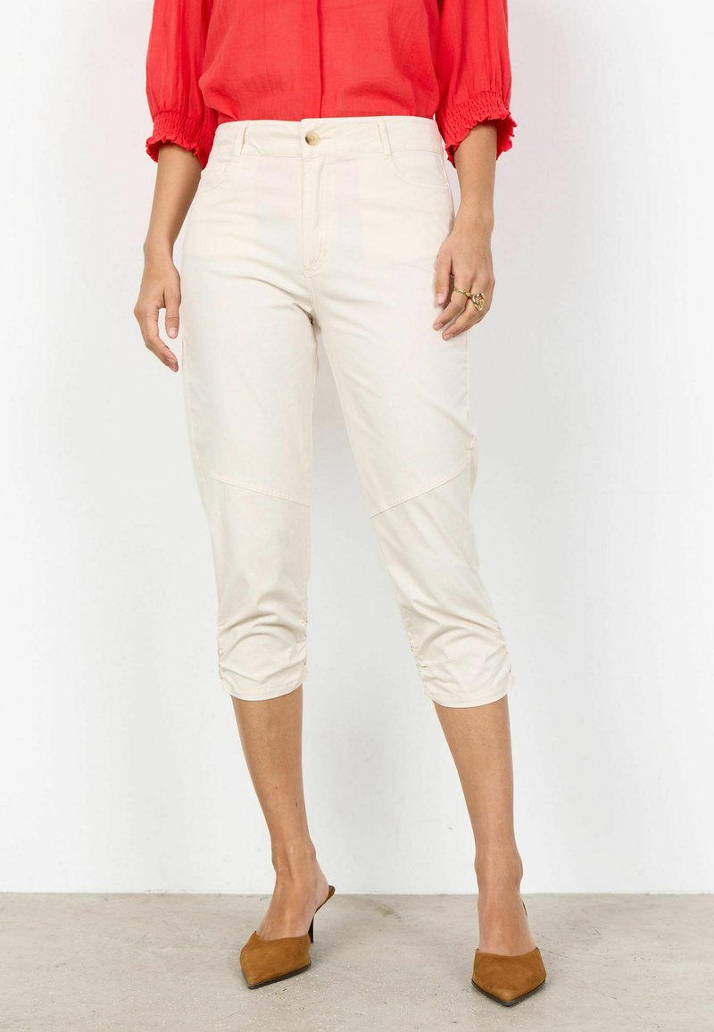 Femme portant un pantalon court écru, un chemisier rouge à manches bouffantes et des mules pointues marron, debout sur un sol neutre.