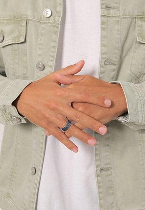 Anello d'argento con una finitura testurizzata e opaca indossato sulle dita, le mani poggiano su una giacca in denim verde chiaro e una camicia bianca sotto.