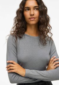 Maglietta grigia a maniche lunghe con texture a coste, scollatura rotonda e design aderente. Indossata con le braccia incrociate, senza motivi o decorazioni visibili.
