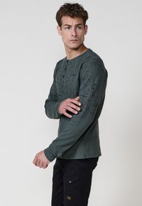 Hosszúujjú henley póló sötétzöld színben, texturált mintázattal és három fekete gombbal a nyaklánál, fekete cargo nadrággal viselve.