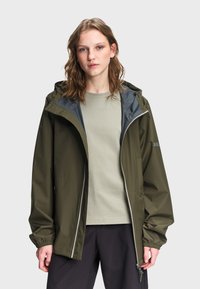 Νεαρό άτομο που φοράει ανοιχτό μπουφάν με κουκούλα σε ελιά πράσινο χρώμα πάνω από ανοιχτό γκρι πουκάμισο και μαύρο παντελόνι, στέκεται μπροστά σε ένα απλό φόντο.