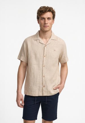 Mand iført en beige kortærmet skorte med knapper og marineblå shorts, stående mod en ensfarvet hvid baggrund.