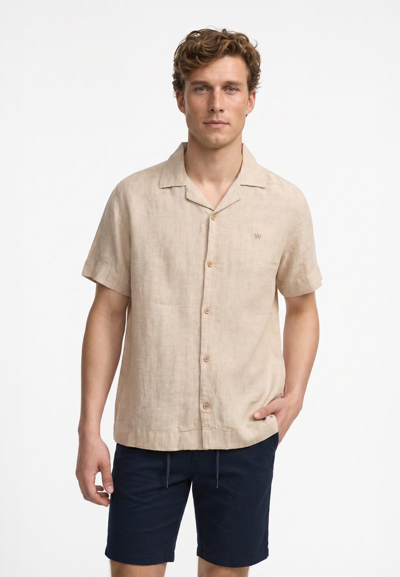 Homme portant une chemise beige à manches courtes boutonnée et un short bleu marine, debout devant un fond blanc uni.