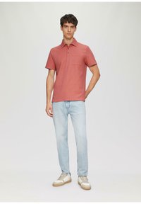 Rot gestreiftes Poloshirt mit Kragen und einer Brusttasche, kombiniert mit hellblauen Jeans und beigen Sneakers, vor einem schlichten Hintergrund stehend.