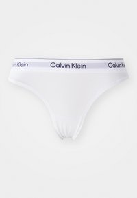 Bijela pamučna tanga gaćice s mekim elastičnim pojasom koji ima plave pruge i otisnutim natpisom "Calvin Klein" u plavoj boji.