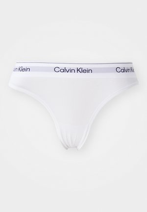 Bijela pamučna tanga gaćice s mekim elastičnim pojasom koji ima plave pruge i otisnutim natpisom "Calvin Klein" u plavoj boji.