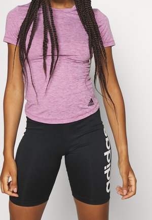 Personne portant un t-shirt Adidas rose ajusté et un short de vélo Adidas noir avec le logo sur la cuisse gauche, debout devant un fond uni.