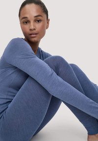 Vrouw zittend met gebogen knieën, gekleed in een blauwe lange mouwtop en bijpassende legging, tegen een effen lichte achtergrond.