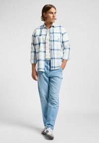 Camisa de cuadros azul claro de botón sobre una camiseta blanca, combinada con jeans azules claros y zapatillas blancas con rayas negras.