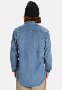 Blauwe corduroy shirt met lange mouwen, een button-down kraag en een rugpasdetail. Heeft een gebogen zoom en knoopmanchetten.