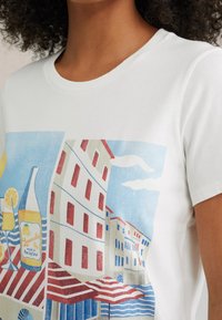 Witte katoenen t-shirt met een kleurrafische afbeelding van gebouwen, gestreepte luifels en een fles met citroen tegen een zeeachtergrond.