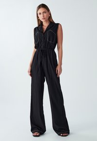 Zwarte mouwloze jumpsuit met een kraag, twee opgestikte zakken aan de voorkant, elastische tailleband en brede pijpen. Zacht, textuurstof met contrasterende stiksels.